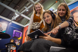 Drei Frauen schauen sich etwas auf einem Tablet an. Eine Frau hat Kopfhörer auf.