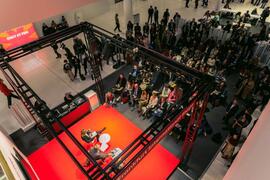 Overhead view of an event area: a red stage framed by a truss structure, surrounded by many visitors standing and seated.