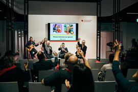 Foto der International Stage mit Zuschauerbereich von einer The Hof Live-Veranstaltung. Personen auf der Bühne und im Zuschauerraum erheben ihre Gläser und sind fröhlich.