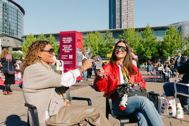 Zwei Besucher*innen sitzen in der Sonne auf dem Freigelände der Frankfurter Buchmesse und stoßen mit ihren Getränken an. 