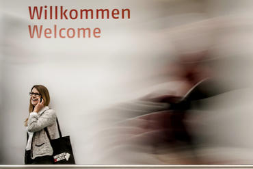 Eine junge Frau steht vor einer Wand, auf der "Willkommen" auf deutsch und englisch steht