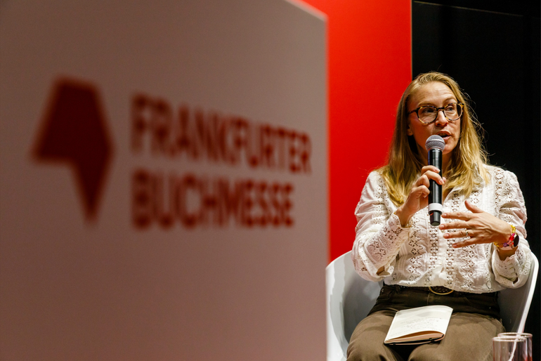 Elisabeth Jaquette speaks into a microphone on stage. The blurred Frankfurter Buchmesse logo is visible in the background.