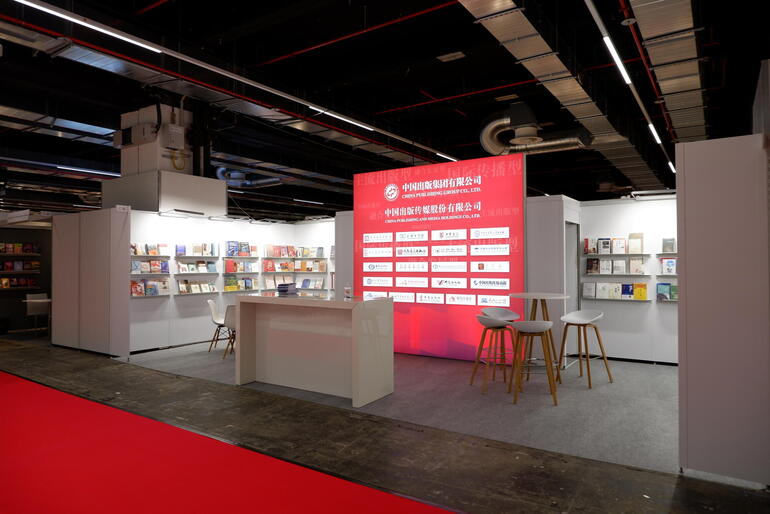 Messestand mit weißer Theke im Vordergrund, weißen Bücherregalen im Hintergrund und einer rot leuchtenden Leinwand in der hinteren Mitte. 