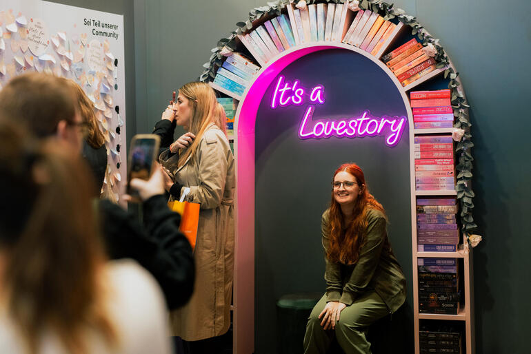 Bücherfans machen Fotos vor einem Bücherregal, auf dem mit Leuchtschrift der Spruch "It's a Lovestory" steht. 