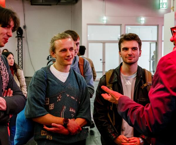 Mehrere junge Erwachsene sprechen bei einer Veranstaltung mit einer älteren Person; im Hintergrund sind weitere Gäste in einem hellen Innenraum zu sehen.