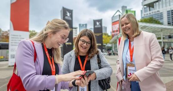 Drei Damen stehen vorm Messeeingang und schauen gemeinsam auf ein Smartphone.