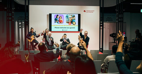 Foto der International Stage mit Zuschauerbereich von einer The Hof Live-Veranstaltung. Personen auf der Bühne und im Zuschauerraum erheben ihre Gläser und sind fröhlich.