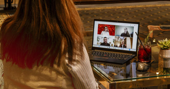 A person sits in a lounge in front of a laptop during a video call; the screen shows four participants in a tiled view, with drinks and a small plant on the glass table beside it.