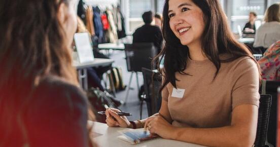 Two women sitting at a desk, talking to each other