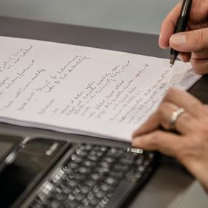Schreibtisch mit Tastatur und Händen, die auf Papier schreiben