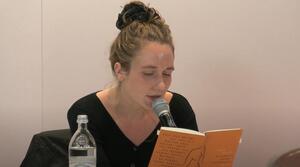 Eine Frau sitzt an einem Tisch und liest aus einem Buch vor, das sie aufgeschlagen vor sich hält. 