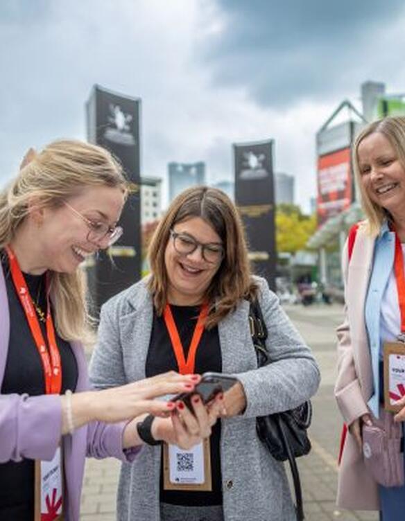 Drei Damen stehen vorm Messeeingang und schauen gemeinsam auf ein Smartphone.