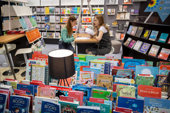 Zwei Personen sitzen an einem Gemeinschaftsstand auf der Frankfurter Buchmesse, umringt von Bücherregalen