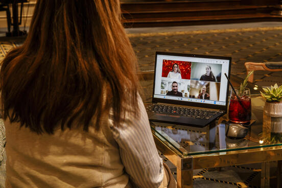 A person sits in a lounge in front of a laptop during a video call; the screen shows four participants in a tiled view, with drinks and a small plant on the glass table beside it.