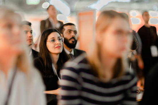 Foto von Menschen, die gemeinsam in einer Messehalle sitzend einem Vortrag lauschen. Die Blicke alle in dieselbe Richtung gerichtet. 