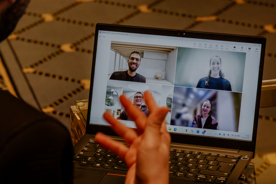Person nimmt an einem Video-Call am Laptop teil; vier Teilnehmende sind im Raster auf dem Bildschirm zu sehen.
