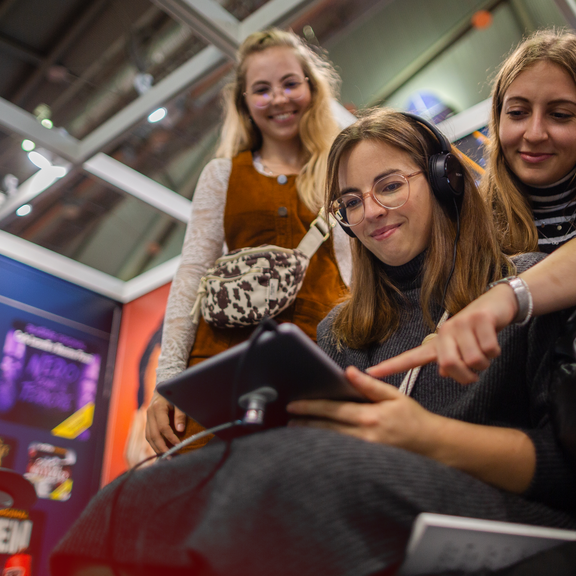 Drei Frauen schauen sich etwas auf einem Tablet an. Eine Frau hat Kopfhörer auf.