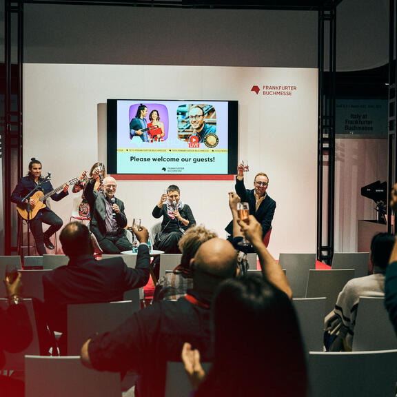 Picture of the International Stage: The speakers on stage and the audience in the public of the live The Hof Event raise their glasses.