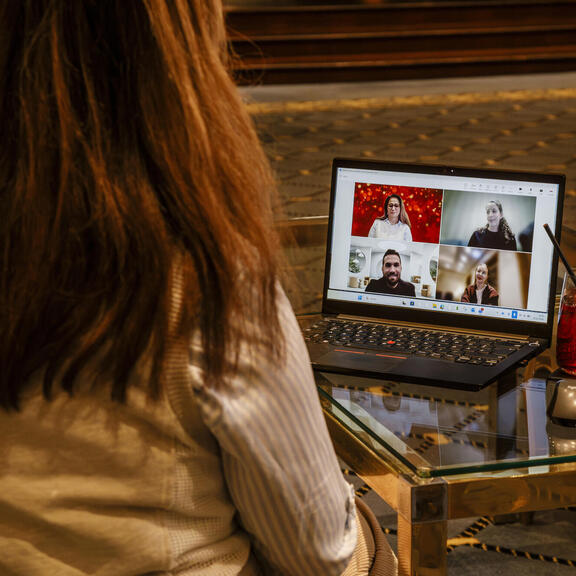 A person sits in a lounge in front of a laptop during a video call; the screen shows four participants in a tiled view, with drinks and a small plant on the glass table beside it.
