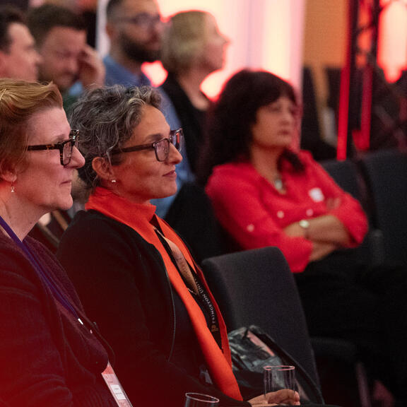 Two attendees sit in the front row of an event space, listening attentively, with more audience members visible in the background under warm, reddish lighting.