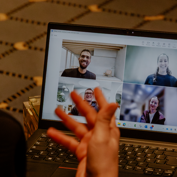 Person nimmt an einem Video-Call am Laptop teil; vier Teilnehmende sind im Raster auf dem Bildschirm zu sehen.