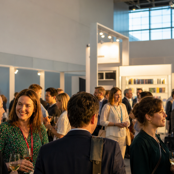 People mingle and talk at a reception in a bright exhibition hall surrounded by bookshelves and trade-fair stands; several attendees are holding wine glasses.