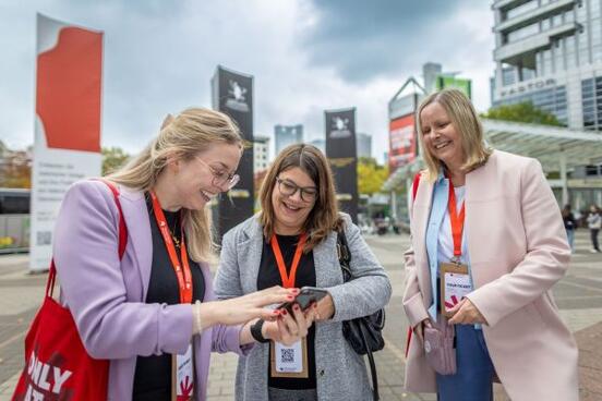 Drei Damen stehen vorm Messeeingang und schauen gemeinsam auf ein Smartphone.