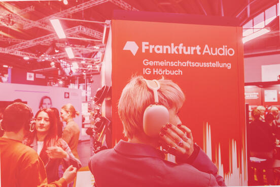 A person wearing large over-ear headphones in front of an orange ‘Frankfurt Audio’ stand at a trade fair, surrounded by visitors