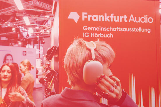 Person mit großen Over-Ear-Kopfhörern vor einem orangefarbenen „Frankfurt Audio“-Stand auf einer Messe, umgeben von Besucher:innen