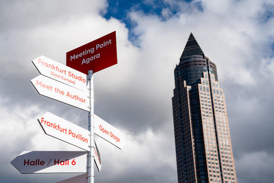 Foto von einem Wegweiser auf der Freifläche des Messegeländes mit Messeturm im Hintergrund. 