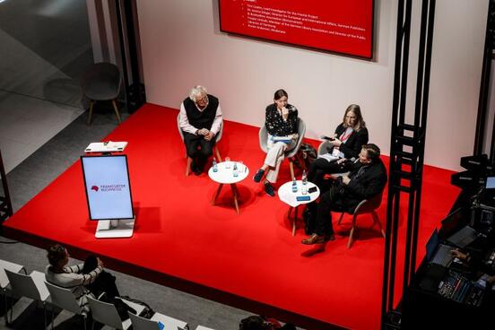 Podiumsdiskussion auf roter Bühne: Vier Personen sitzen in einer Runde mit kleinen Tischen; davor sind einzelne Stuhlreihen zu sehen.