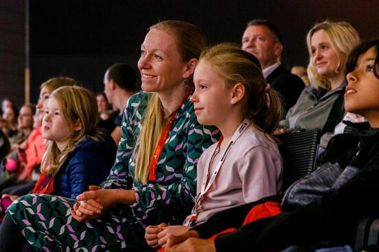 Publikum in einem Saal: Eine Frau und zwei Kinder sitzen in der ersten Reihe und schauen aufmerksam zur Bühne.