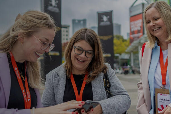 Drei Messegäste mit Namensausweisen stehen im Freien vor Messe-Beschilderung und schauen gemeinsam auf ein Smartphone.