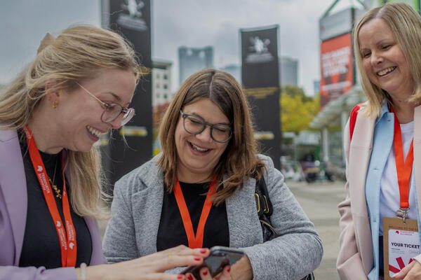 Drei Messegäste mit Namensausweisen stehen im Freien vor Messe-Beschilderung und schauen gemeinsam auf ein Smartphone.